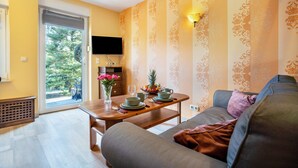 Una sala de estar con un sofá, una mesa de comedor dispuesta con platos y tazas, un televisor en la pared y un ventanal con vistas a zonas verdes.