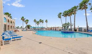 A pool area with blue lounge chairs, palm trees, and a building with a blue awning. - LIV Sea Cottage, Boat dock, Pool, Golf! (Port Isabel)
