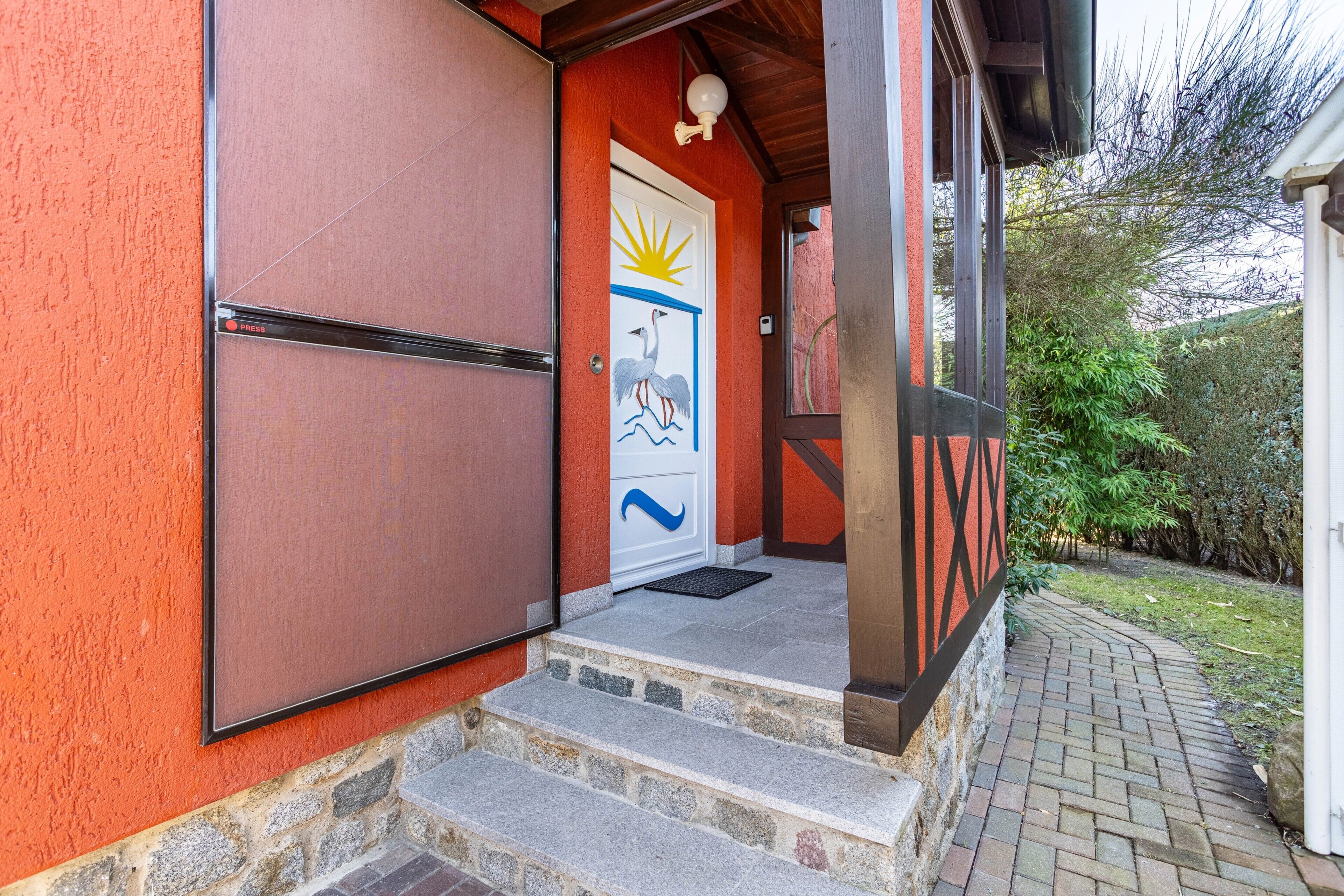 Una entrada de edificio con un muro rojo, una puerta blanca con diseño de sol y pájaro, y escalones de piedra que conducen a un porche.