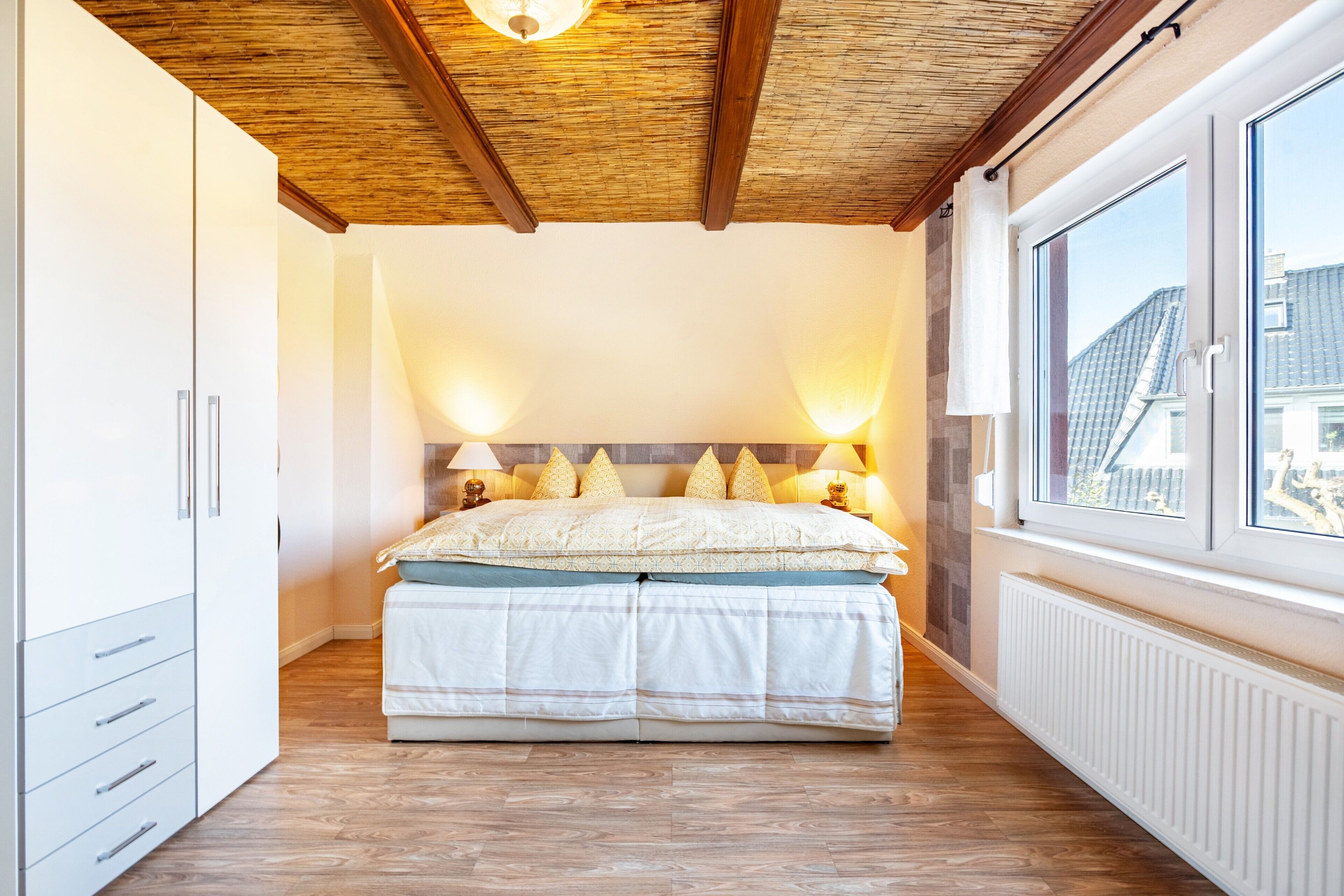 A bedroom with a wooden ceiling, a bed with white bedding, two bedside lamps, and a window with a view of buildings.