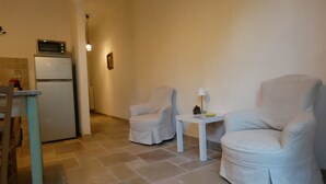 Un espacio habitable compacto con kitchenette, dos sillones blancos, una mesita auxiliar y una lámpara.