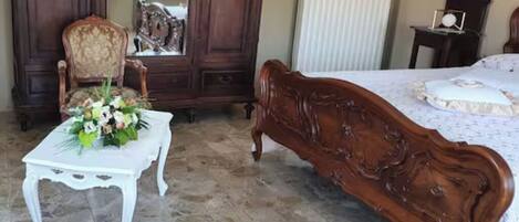 A bedroom with a wooden bed, a white table with a floral arrangement, a wooden wardrobe, and a chair.