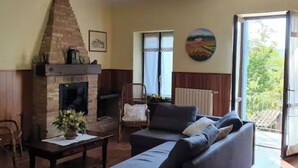 A living room with a stone fireplace, a sofa, a coffee table, and a chair. There is a window with a view of greenery outside. - CASA MILU' (Piemonte)