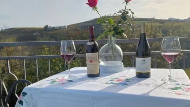 A table with a floral tablecloth, two wine bottles, and two glasses of wine on a balcony with a scenic view. - CASA MILU' (Piemonte)