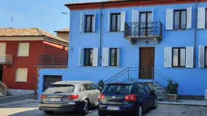 A two-story building with a blue facade and white shutters. There are two cars parked in front. - CASA MILU' (Piemonte)