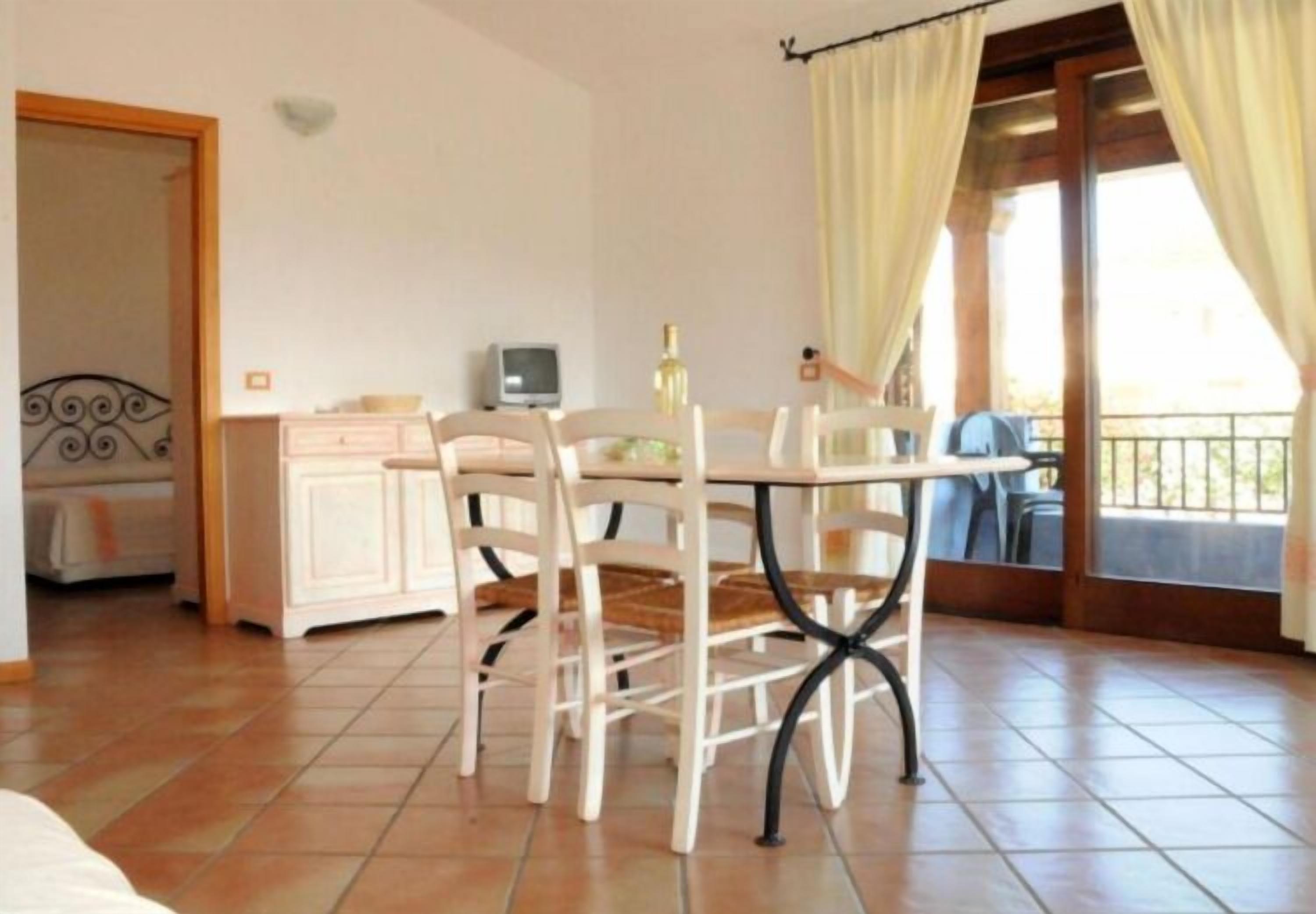 Une pièce comprenant une table à manger, des chaises, un petit meuble, un téléviseur et un balcon avec des rideaux.