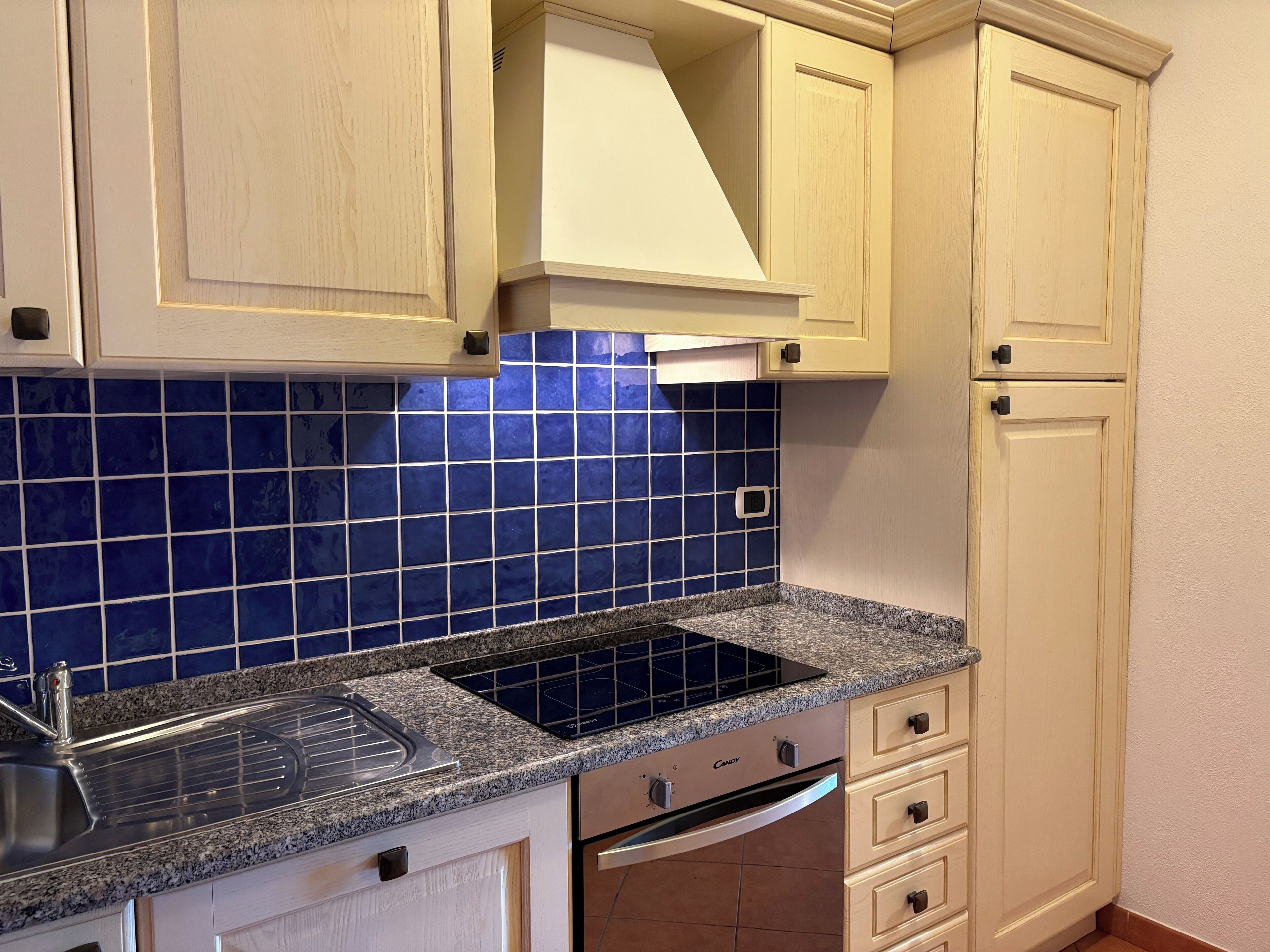 Une cuisine avec des meubles de couleur bois clair, un dosseret de carrelage bleu, un plan de travail en granit, un four encastré et un évier en acier inoxydable.