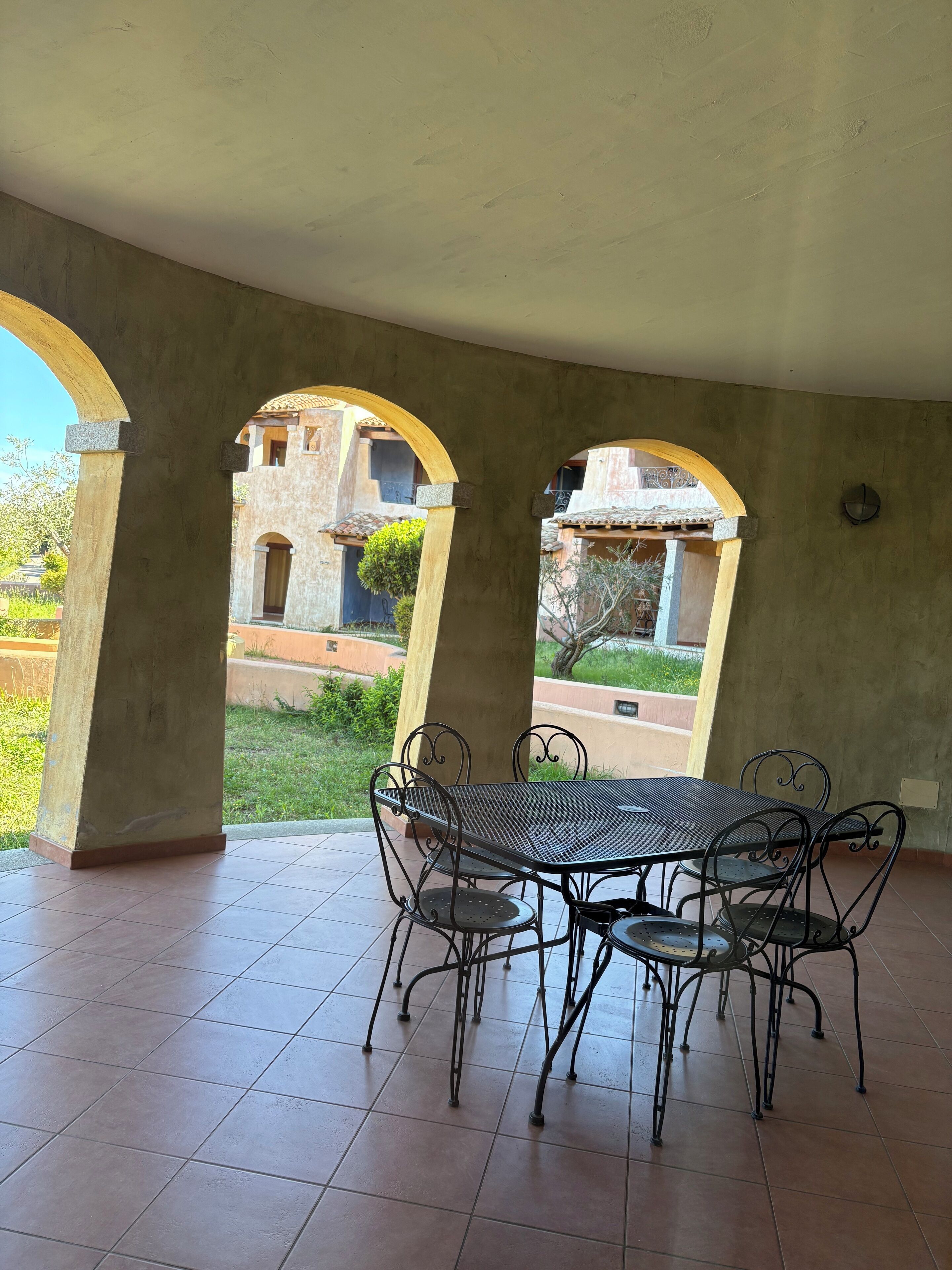 Ein möblierter Patio-Bereich mit Tisch und Stühlen, umgeben von Bögen und Blick auf ein Gebäude mit Ziegeldach.