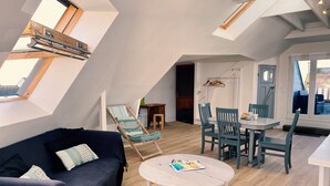 Et loftstudie med højloftet rum, ovenlys, en mørkeblå sofa, et hvidt sofabord, samt et spiseområde med blå stole og et hvidt bord.
