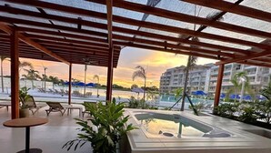 Un espacio al aire libre techado con sillones de descanso, un jacuzzi y vistas a un hotel y una playa al atardecer.