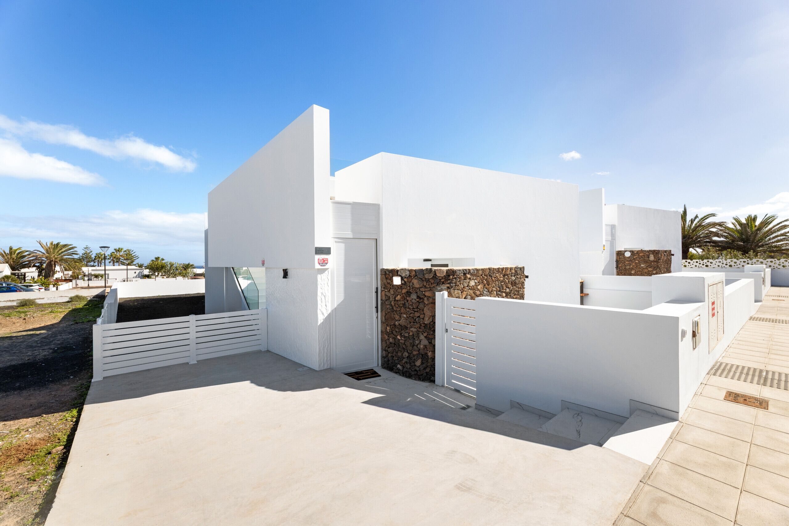 Un bâtiment moderne de couleur blanche, avec une entrée surmontée d’un mur en pierre, une allée bordée de portails et un ciel dégagé.