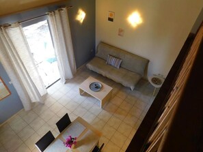 A living room with a sofa, a coffee table, and a dining table with chairs. There are curtains on the window, and wall-mounted lights.