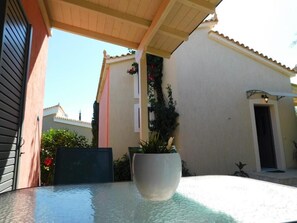 A glass-top table with a white pot in the center, a wooden pergola overhead, and a white building with a tiled roof in the background.