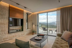 A modern living room with a stone wall, a flat-screen TV, a sofa, a coffee table, and a balcony with a view. - Villa Phos, Sea view, Luxury, private pool, BBQ (Τοπική Κοινότητα Καλλεργιανών)