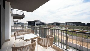 A balcony with wicker furniture and a glass table, overlooking a modern cityscape with buildings and a road. - Stylish 3BR Serviced Apart At Cairo Festival City (Cairo Governorate)