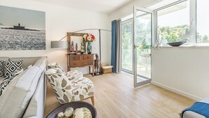 Een woonkamer met een bank, een houten dressoir, een vaas met bloemen en een groot raam met uitzicht op groen.