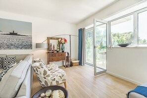 Ein Wohnzimmer mit einer Couch, einem Holzschrank, einer Blumenvase und einem großen Fenster mit Blick auf Grünflächen.
