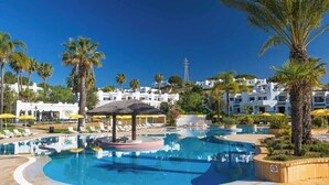 Ein Pool mit Liegestühlen, Palmen und weißen Gebäuden im Hintergrund.