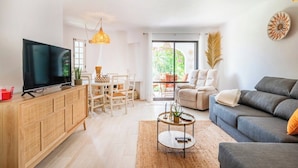 Ein modernes Wohnzimmer mit Flachbildfernseher, Holzschrank, grauem Sofa und Essbereich mit Tisch und Stühlen.