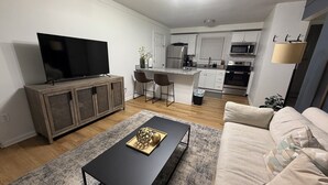 Un salon moderne avec un téléviseur à écran plat, une table basse sombre et un canapé beige. On aperçoit, au fond, une cuisine avec des meubles blancs et des appareils électroménagers en acier inoxydable.