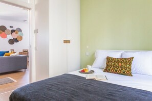 Ein Schlafzimmer mit einem Bett, einem Nachttisch mit einem Buch und einer Fruchtplatte, einem Sofa im angrenzenden Raum und einer Wand mit runder Wandkunst.