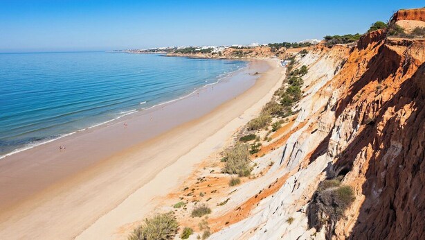 Una playa costera con cielo despejado, arena dorada y un acantilado rocoso.