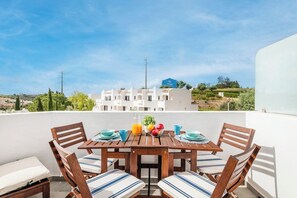 Eine Terrasse mit einem Holztisch, gedeckt für ein Essen, umgeben von Stühlen mit gestreiften Kissen und Blick auf weiße Gebäude im Hintergrund.