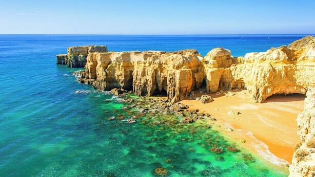 Paisagem costeira com falésias rochosas, água turquesa límpida e uma praia de areia.