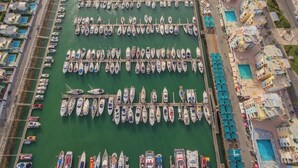 Vista aérea de um porto com vários barcos atracados em fileiras, próximo a um edifício com várias varandas e uma piscina.