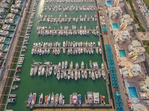 Eine Luftaufnahme eines Hafens mit zahlreichen in Reih und Glied liegenden Booten, neben einem Gebäude mit mehreren Balkonen und einem Schwimmbad.