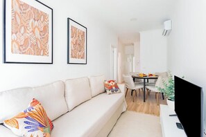 Ein Wohnzimmer mit Sofa, einem Esstisch mit Stühlen, einem Flachbildfernseher und gerahmten Kunstwerken an der Wand.