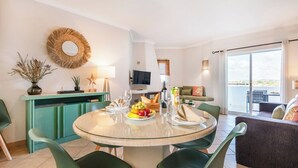 Una sala de estar moderna con una mesa redonda de vidrio, sillas verdes, un sofá, televisión y un balcón con vistas.