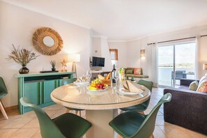 Ein modernes Wohnzimmer mit einem runden Glastisch, grünen Sesseln, einer Couch, einem Fernseher und einem Balkon mit Aussicht.