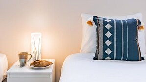 Quarto de hotel com uma cama, uma mesinha de cabeceira com uma xícara e um prato de comida, e uma janela com vista para o verde.