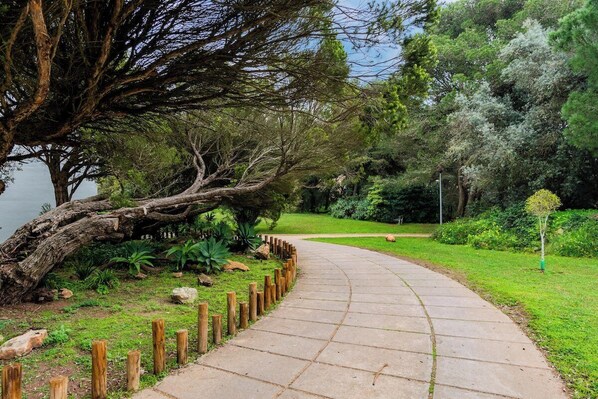Ein gewundener Weg in einem Park, links ein großer Baum und eine Umzäunung entlang des Pfades.