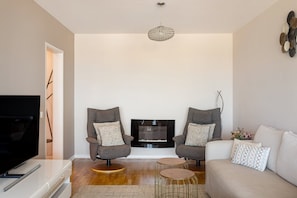 A modern living room with two armchairs, a sofa, a flat-screen TV, a fireplace, and a decorative ceiling light. - Cascais Prime 2BR with Balcony & Sea View (Cascais)