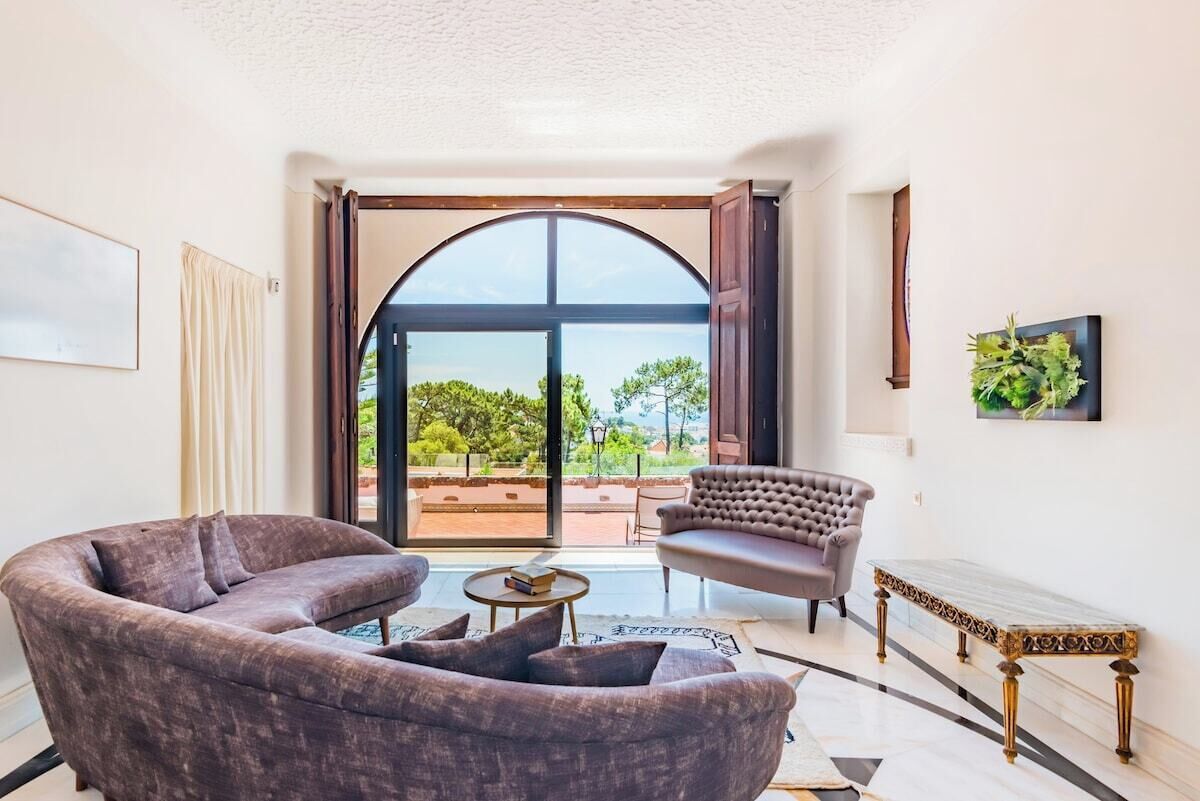 Un salon comprenant un canapé, un fauteuil et une petite table. Une grande fenêtre en arc offre une vue sur la verdure à l’extérieur.