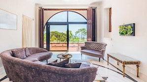 Ein Wohnzimmer mit Sofa, Sessel und einem kleinen Tisch. Ein großes Bogenfenster bietet Blick auf Grünflächen.