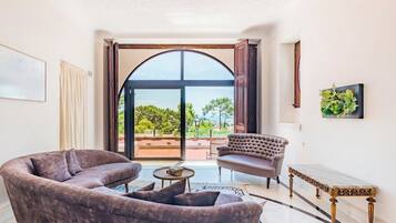 Un salon comprenant un canapé, un fauteuil et une petite table. Une grande fenêtre en arc offre une vue sur la verdure à l’extérieur.