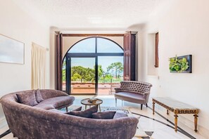 Ein Wohnzimmer mit Sofa, Sessel und einem kleinen Tisch. Ein großes Bogenfenster bietet Blick auf Grünflächen.