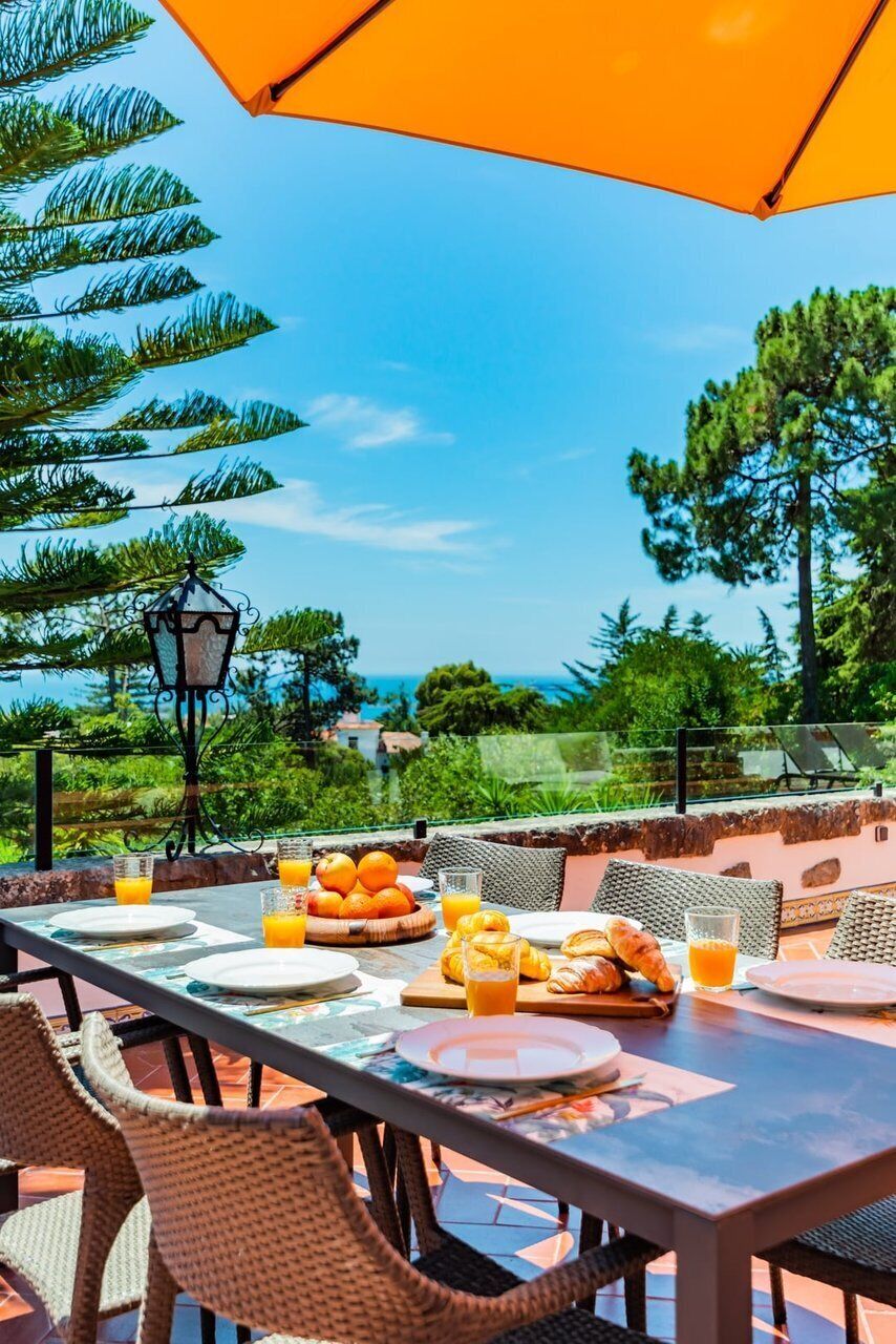 Een terras met een ontbijttafel, compleet met croissants en sinaasappelsap, onder een grote oranje parasol.