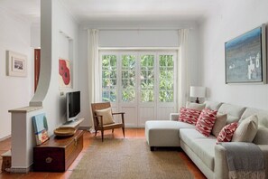 Ein Wohnzimmer mit einer Couch, einem Fernseher, einem Sessel, einem Fenster mit Vorhängen und Kunstwerken an den Wänden.