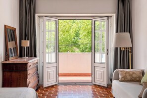 Ein Zimmer mit einem Holzkoffer, einem Bett, einer Lampe und Blick durch eine offene Tür auf einen Balkon.