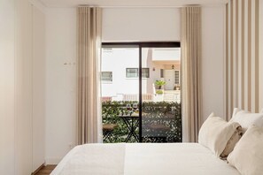 Ein Schlafzimmer mit Bett, Vorhängen, Blick auf einen Balkon mit Pflanzen und einem Tisch mit Weingläsern.