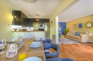 Ein Essbereich mit Holztisch, gedeckt für vier Personen, eine Küche mit dunklen Schränken und Geräten sowie ein Wohnbereich mit Sofa und dekorativen Spiegeln.