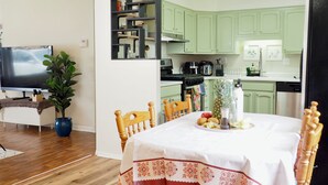 A dining area with a table set for two, a television, a potted plant, and a kitchen in the background.
