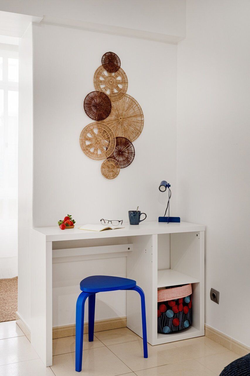 A white desk with a blue chair, a wall-mounted decorative basket, and a small shelf with a lamp.