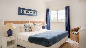 Ein Schlafzimmer mit einem Bett, zwei Nachttischen, einer Nachttischlampe, einem blauen Kunstwerk, einem Korbstuhl und Blick auf die Stadt durch das Fenster.