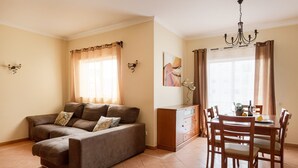 A living room with a sofa, a dining table with chairs, a chandelier, and a window with curtains. - Central Albufeira Stay | Balcony & AC (Albufeira)