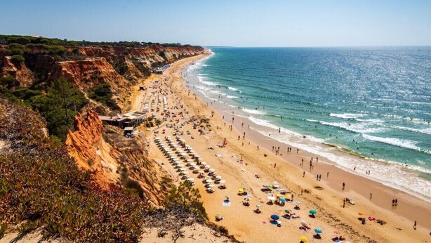 A coastal beach with a long stretch of beach umbrellas and sun loungers, a clear sky, and a rocky cliffside. - Central Albufeira Stay | Balcony & AC (Albufeira)
